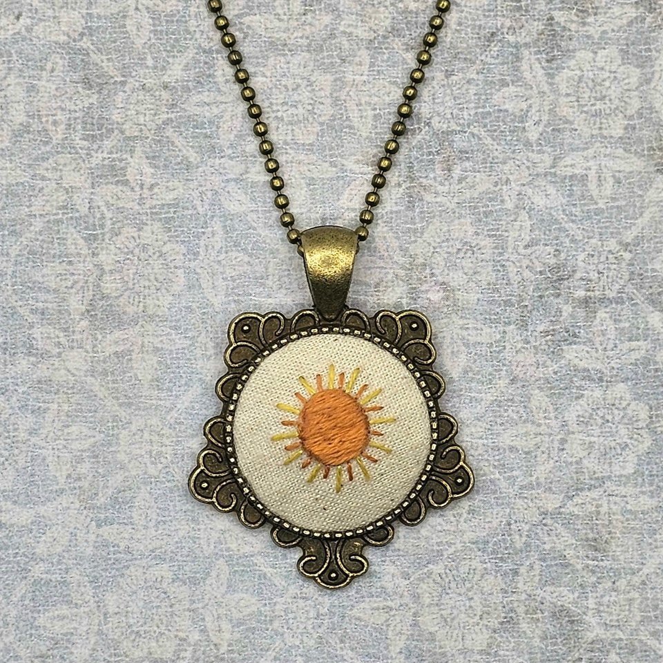 Hand embroidered sunshine necklace in a brass pendant setting centered in a neutral background.