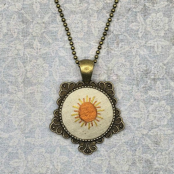Hand embroidered sunshine necklace in a brass pendant setting centered in a neutral background.
